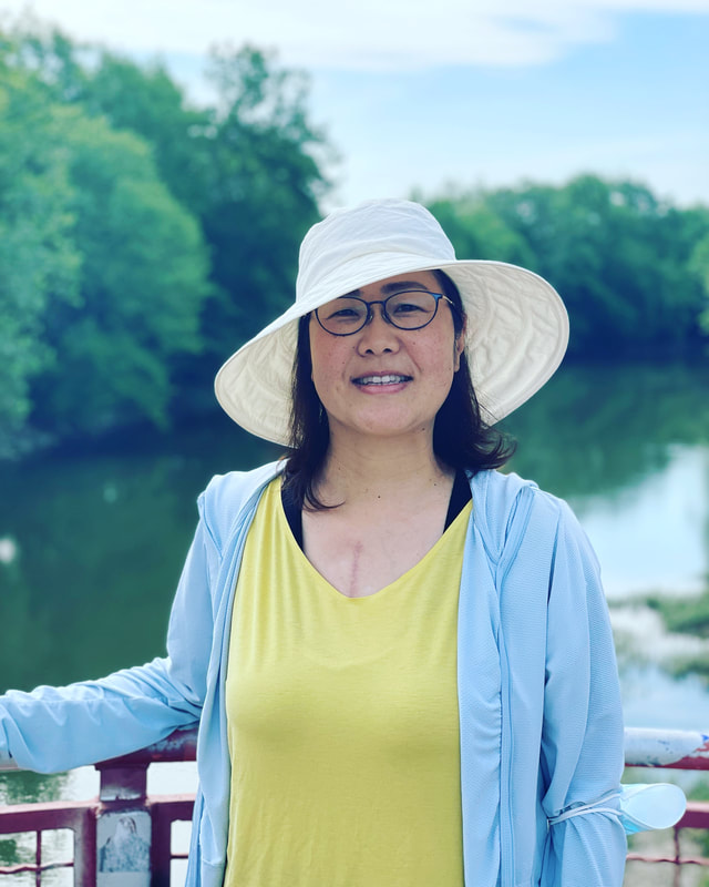Portrait of Motoko outdoors by a river, wearing a white sun hat and a warm smile